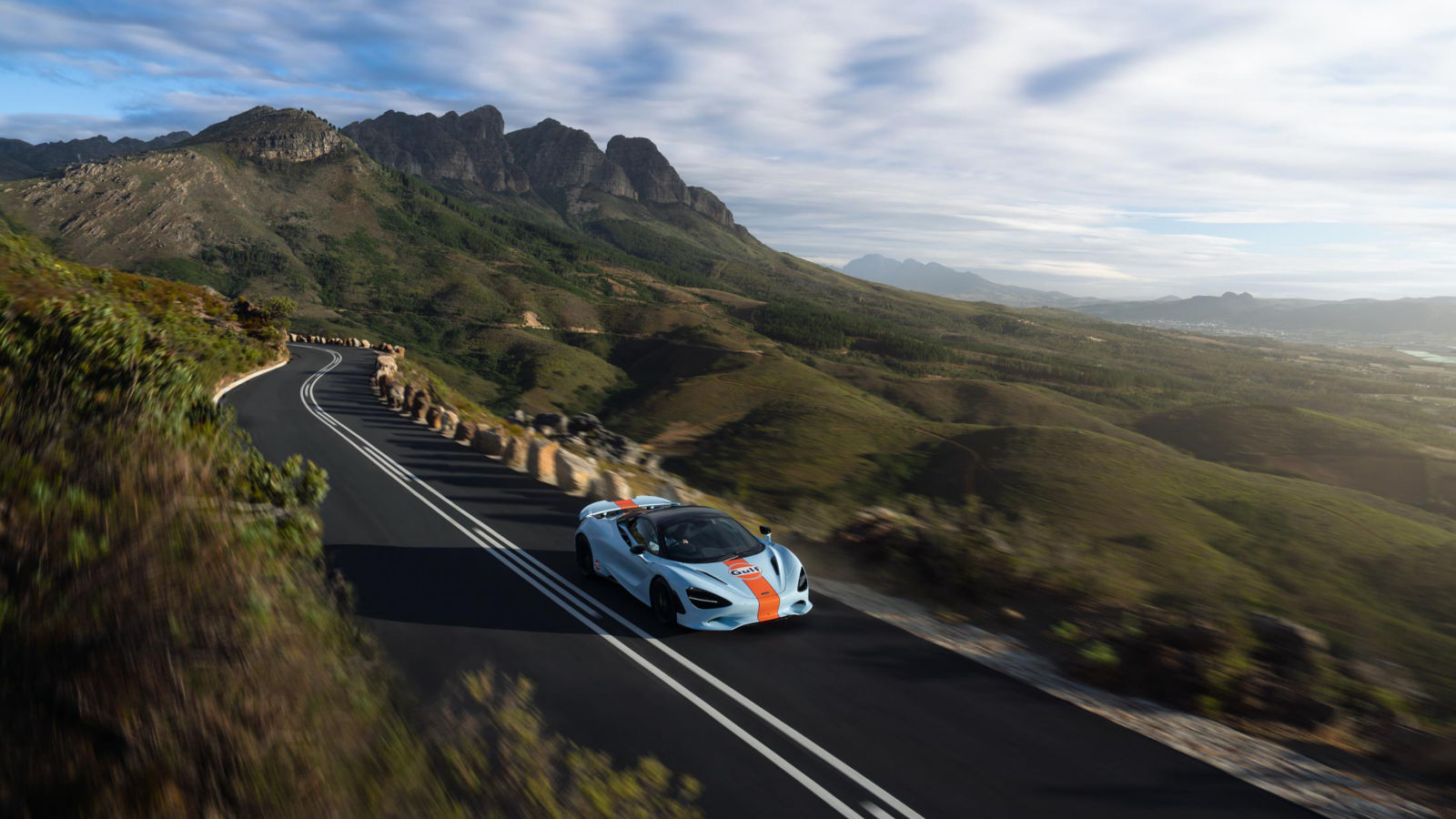 The McLaren and Gulf collab supercar driving at speed along a cliff top with hills in the background.