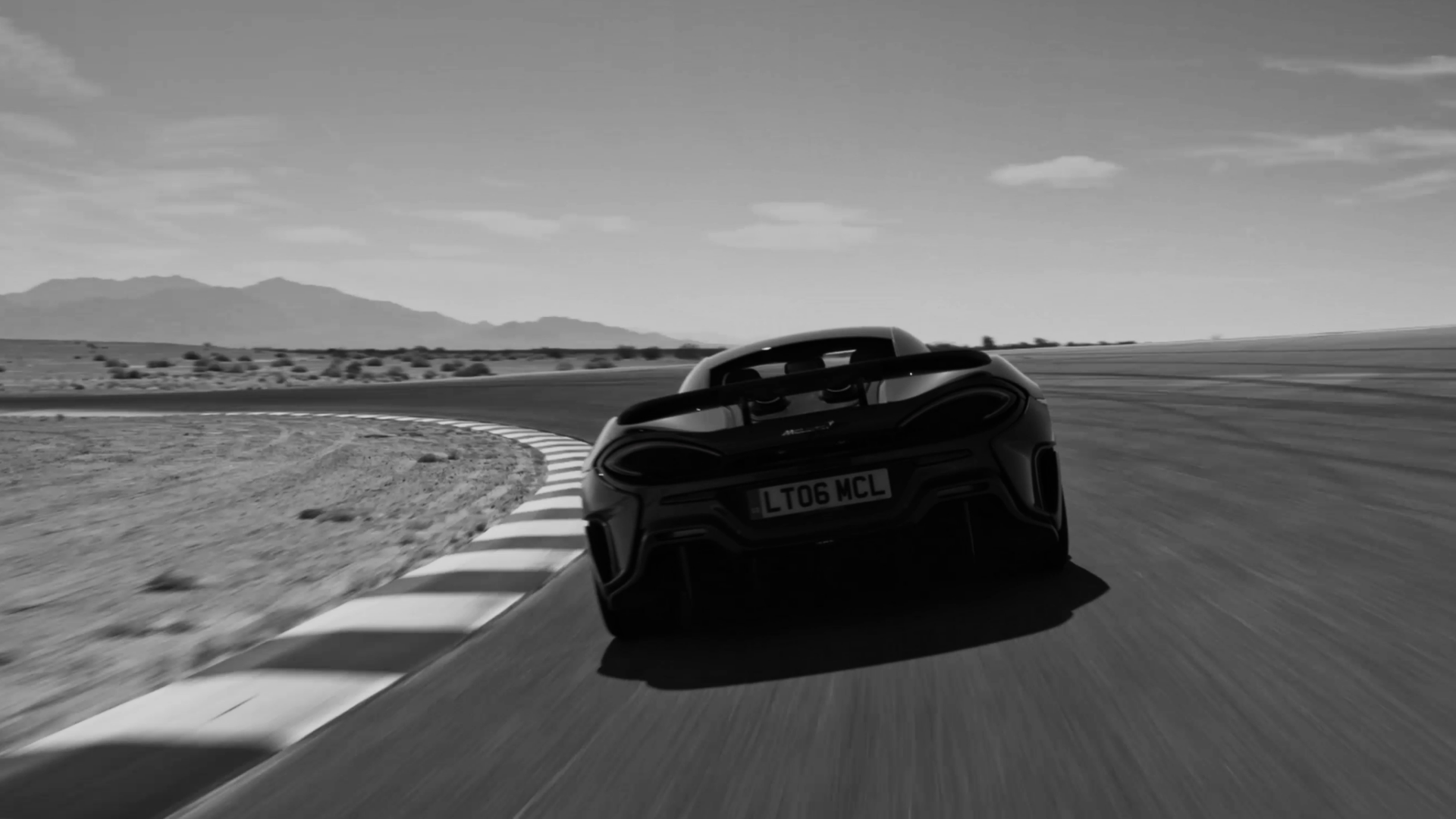 Black and white image of a McLaren LT supercar on a racetrack, captured mid-corner with an active rear wing, emphasizing speed.