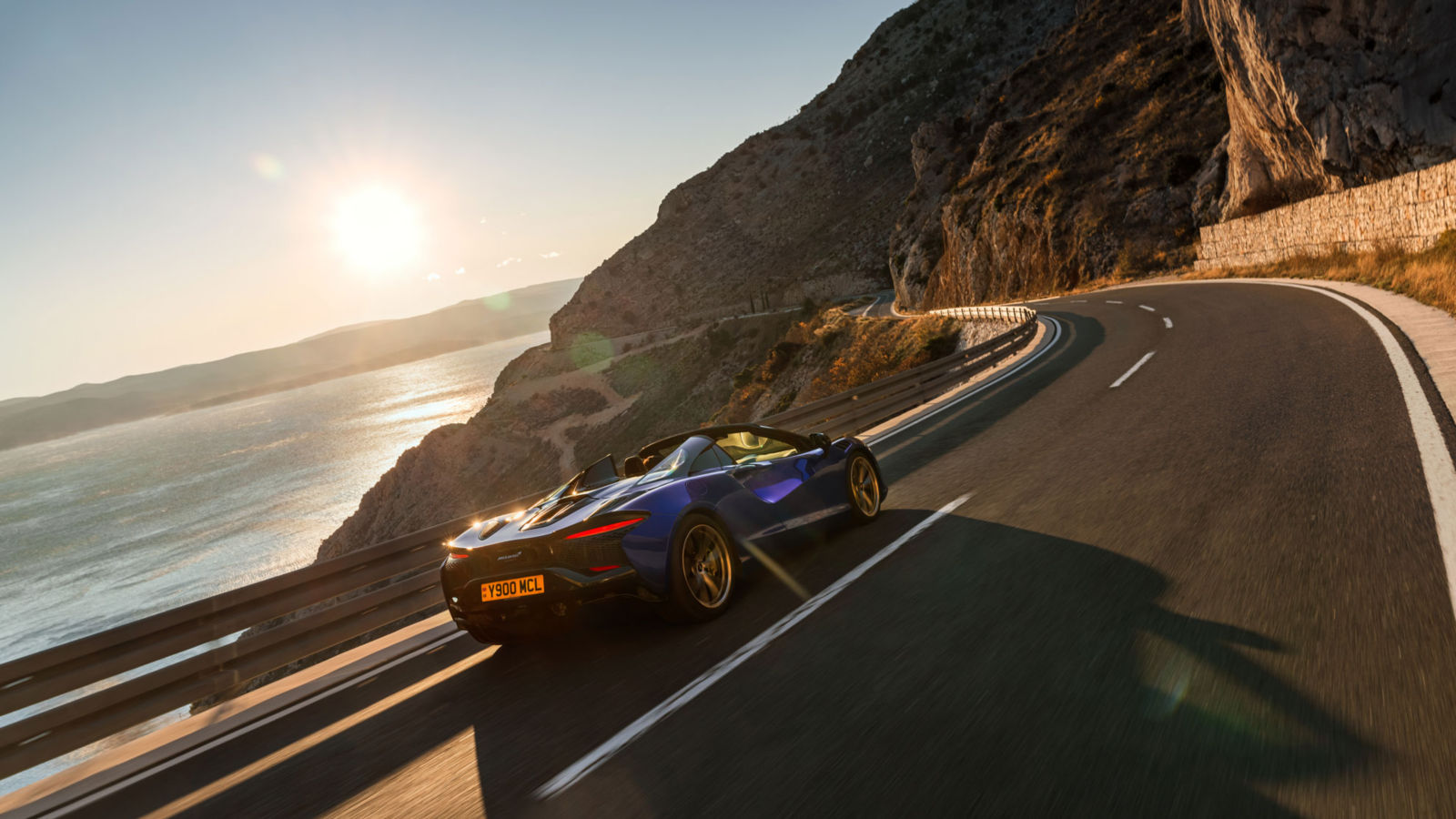 View of the McLaren Artura driving along a cliff top road with the sun setting in the background.