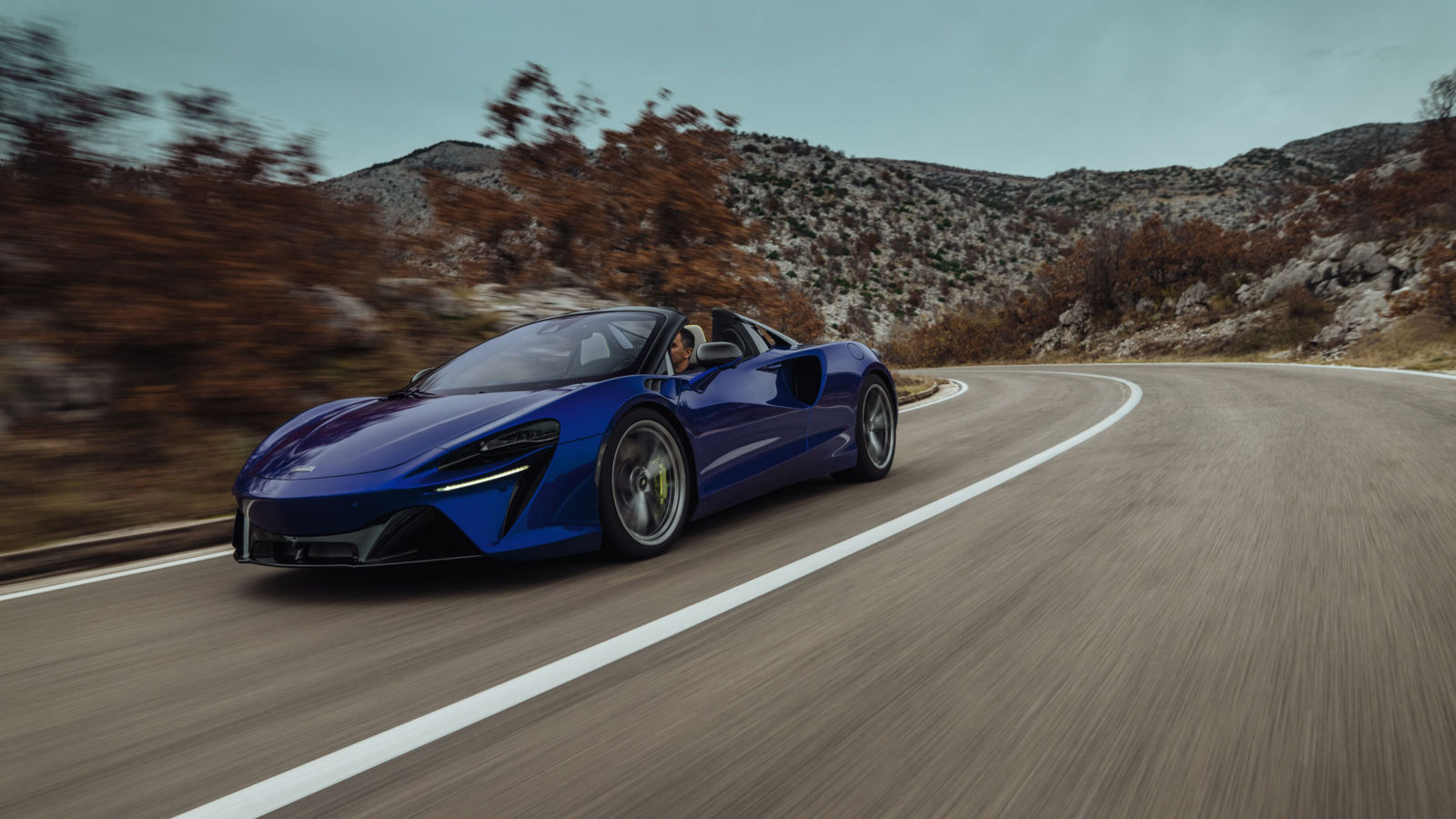 The Mclaren Artura driving at speed around a corner with large hills in the background.