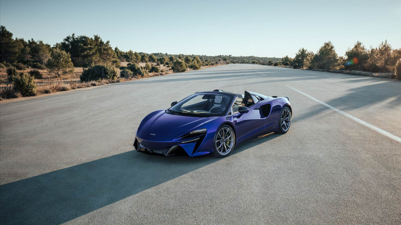 The Mclaren Artura parked in the centre of the road with trees and greenery running alongside either side of the road.