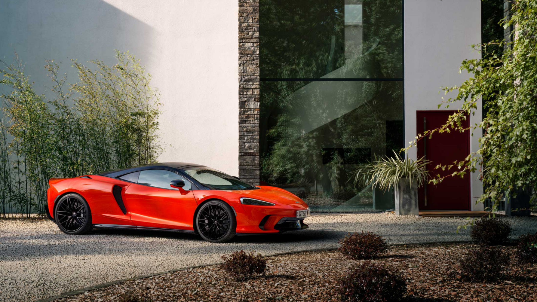 Landscape shot of the offside of the McLaren GTS in red parked on a stoney driveway in front of a grand house.