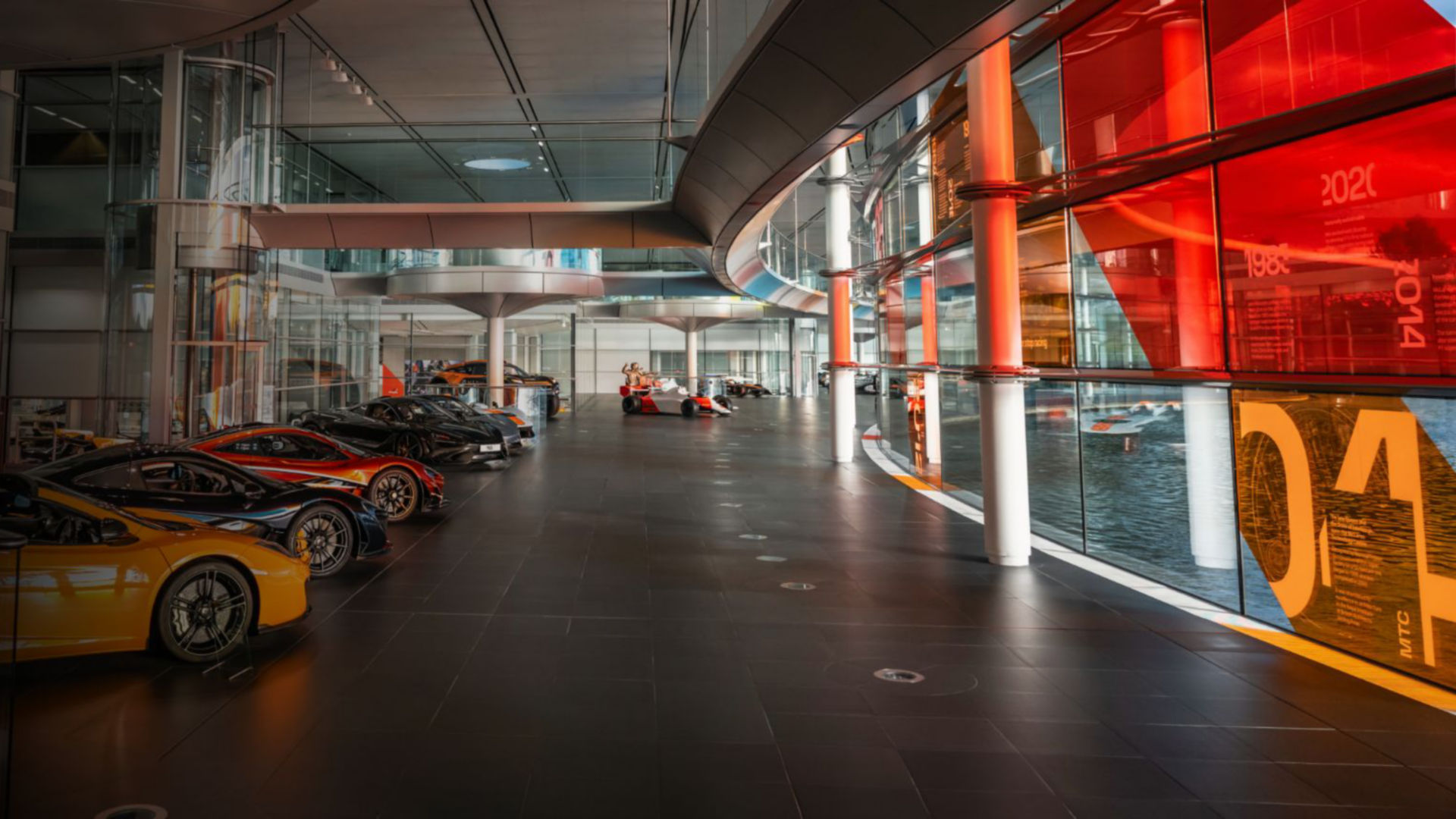 Inside the McLaren headquarters. On the left there is a row of McLaren cars lined up on display. Then to the left there are floor to ceiling windows that overlook the McLaren lake.