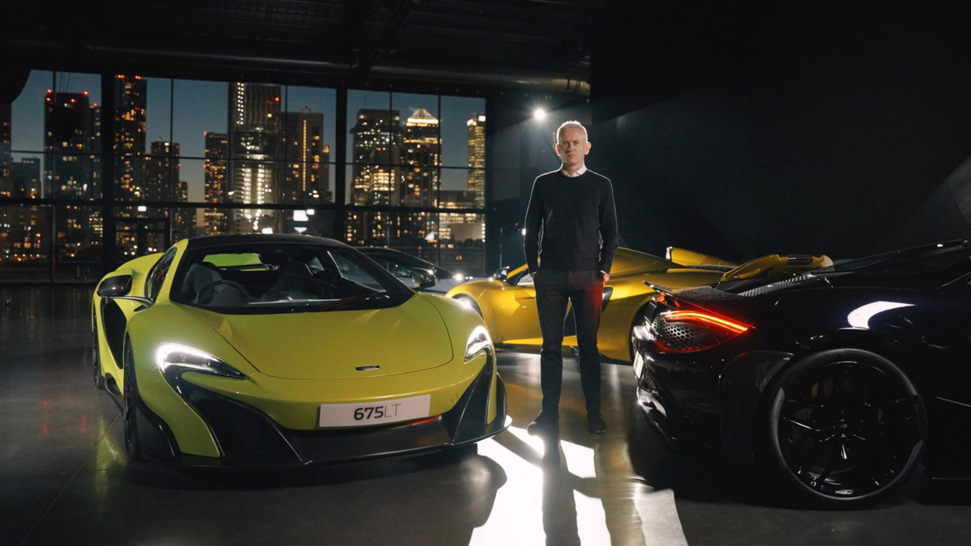 McLaren executive stands among 675LT and other LT models in a dimly lit showroom with a city skyline backdrop.