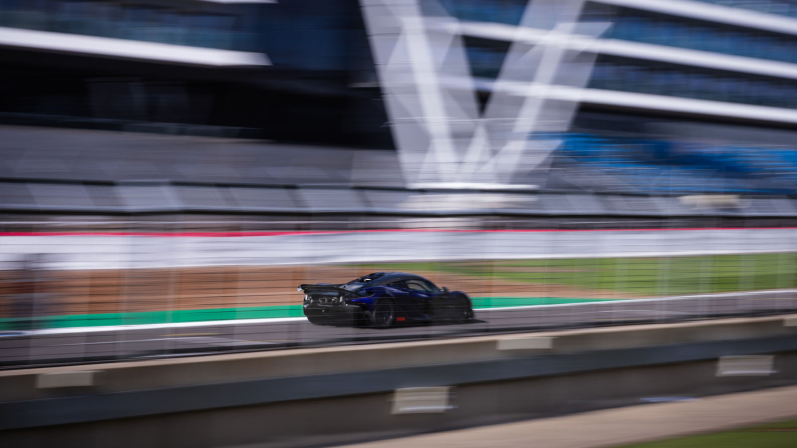 A shot of the rear side of a McLaren W1 prototype in royal blue driving at speed around the Silverstone track with motion blur to enhance the idea of speed.